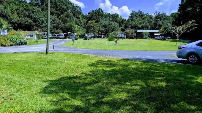 A grassy park area with a curved paved road, several trees, parked cars, and small buildings or trailers in the background under a blue sky with scattered clouds.