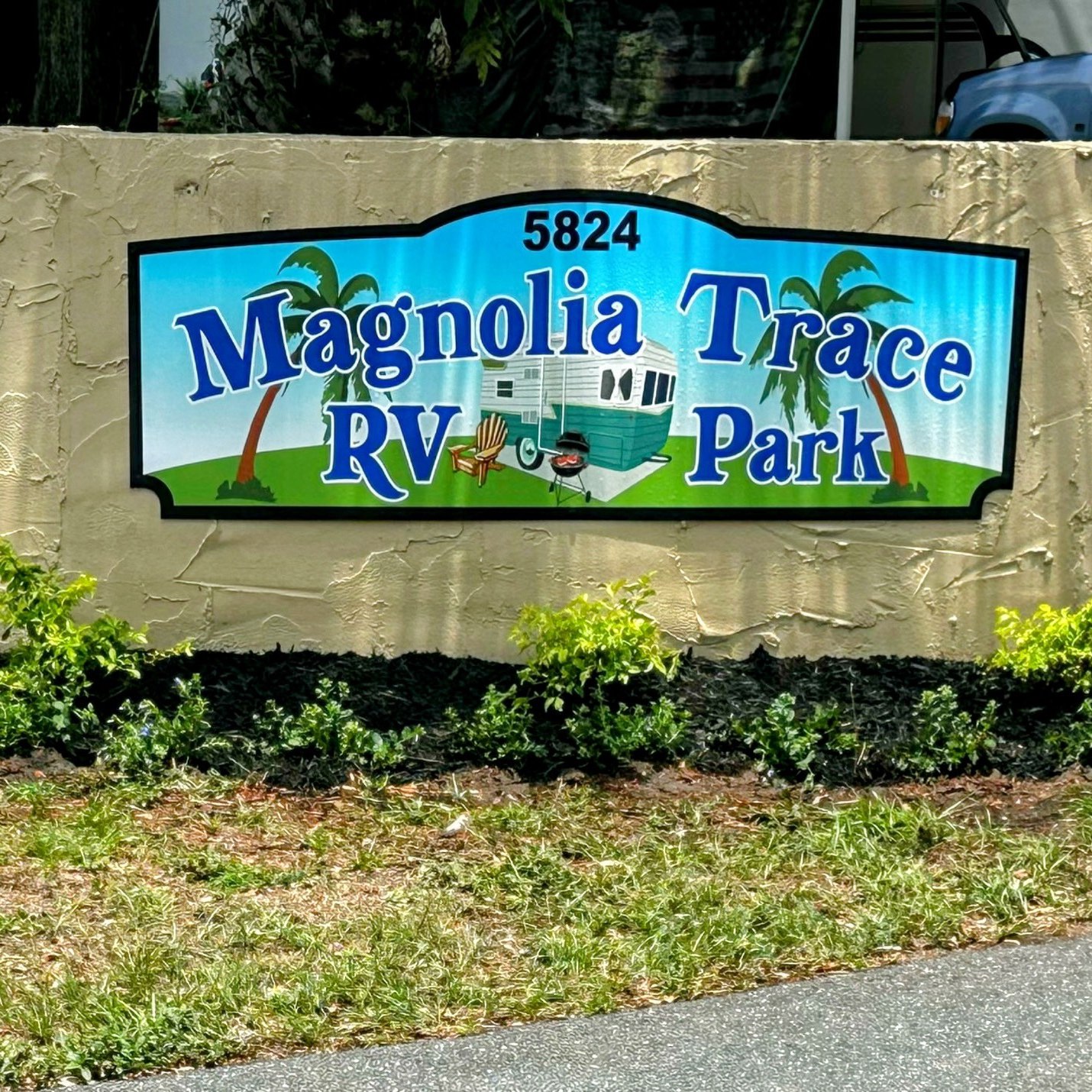 A sign on a beige wall reads “5824 Magnolia Trace RV Park,” with a drawing of an RV, palm trees, chairs, and a campfire on grass. Shrubs and a paved road are in front of the wall.