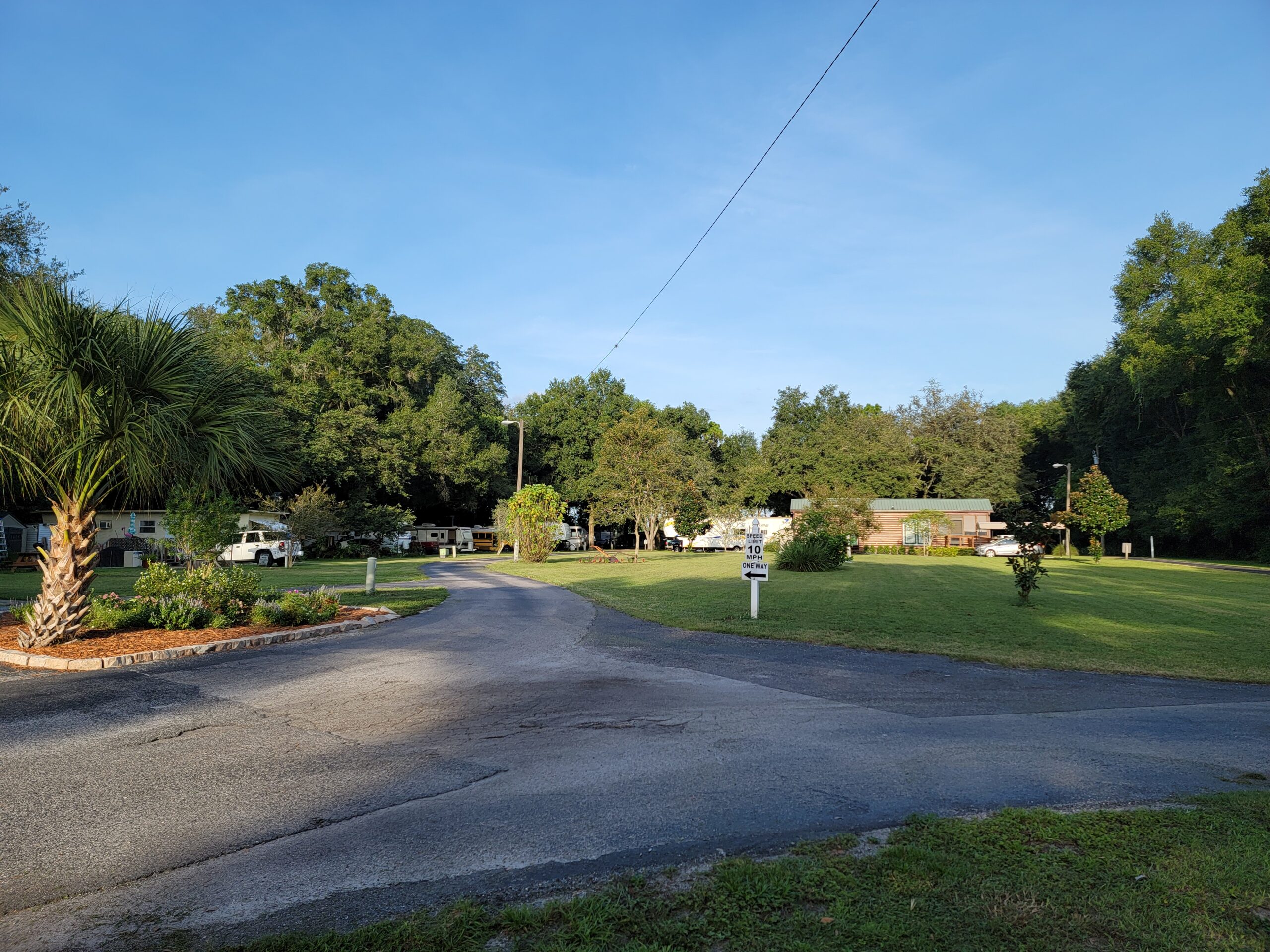A paved driveway curves through a grassy area with scattered trees, parked RVs, and a few small buildings under a clear blue sky. A palm tree is visible on the left side of the image.