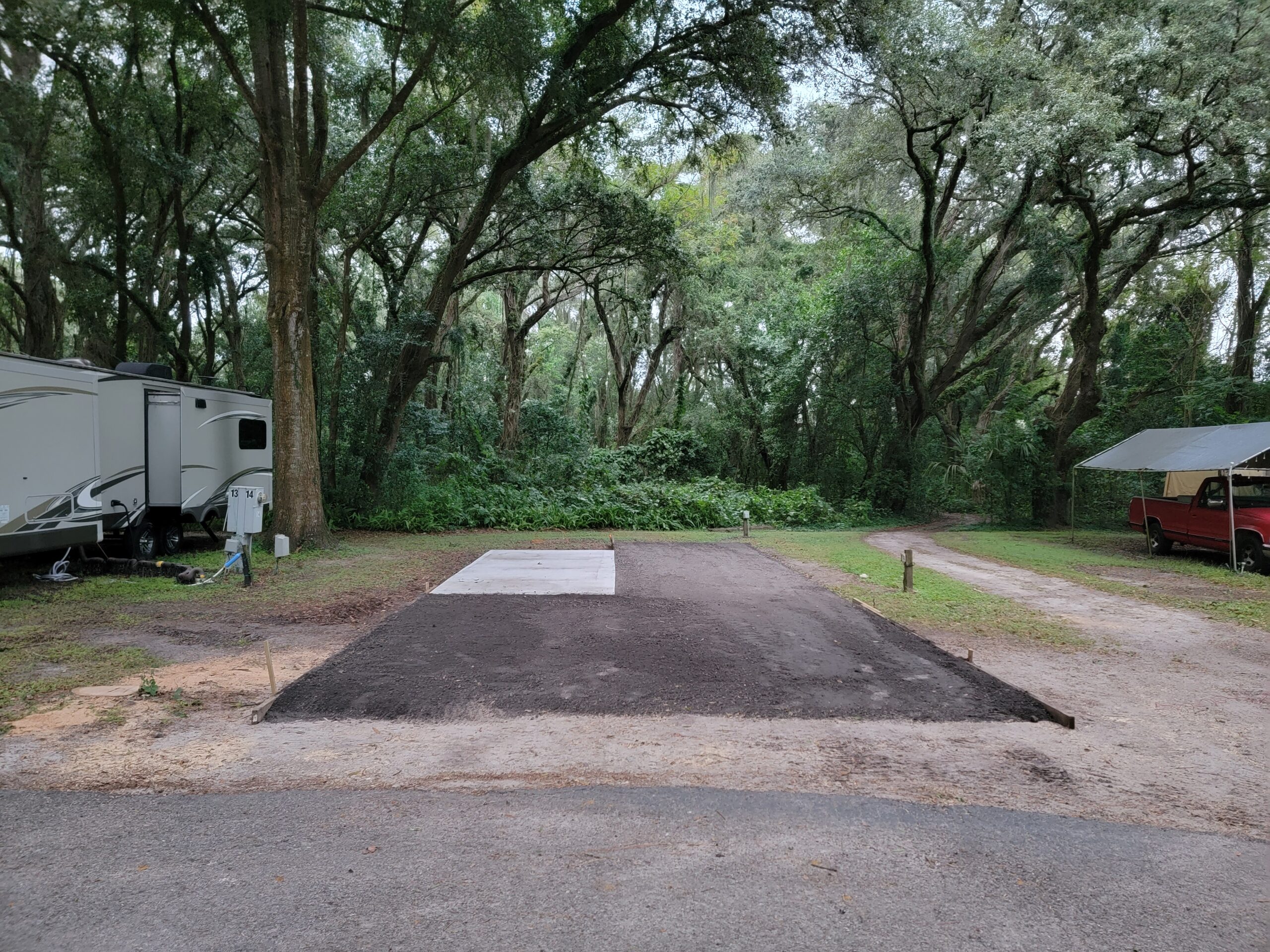 A campsite with a fresh, rectangular patch of leveled soil and a small concrete slab in the center, surrounded by trees. An RV is parked to the left, and a red truck is on the right near a gravel road.